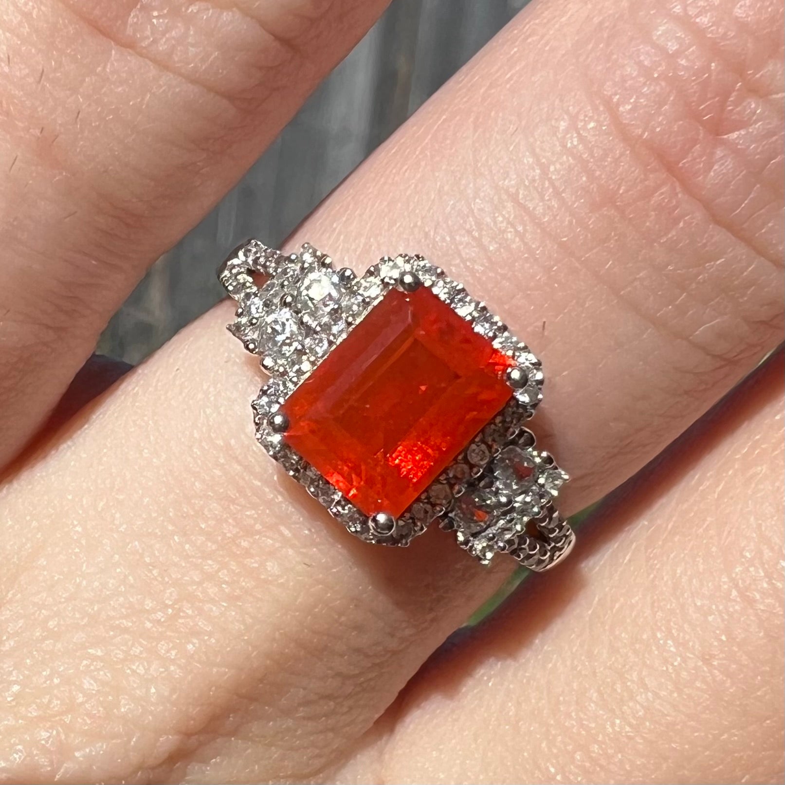 Close-up macro view of a white gold emerald cut fire opal and zircon halo ring worn on a finger in natural light.