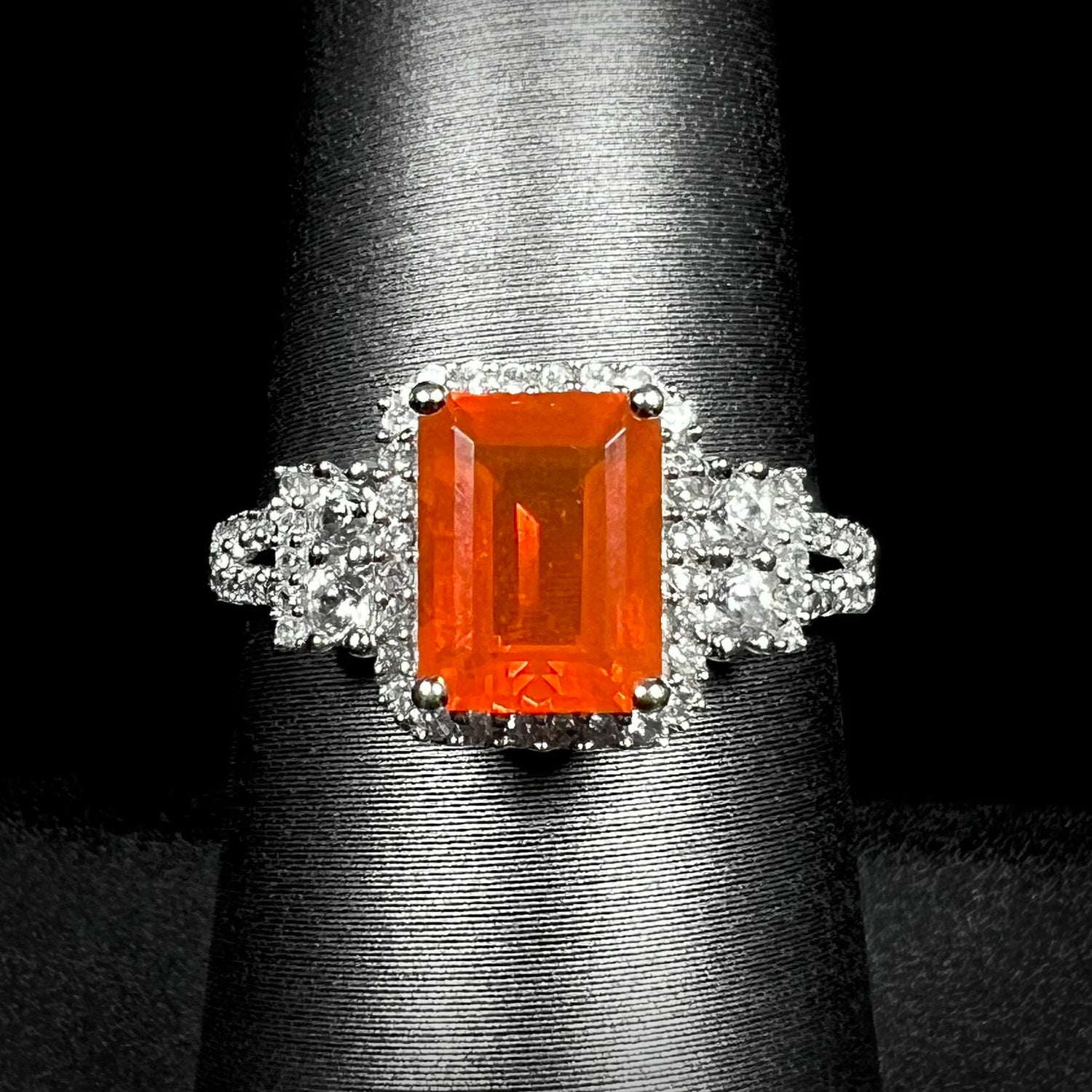 Macro shot of a white gold halo ring set with an orange emerald cut Mexican fire opal on a ring display against a black background.