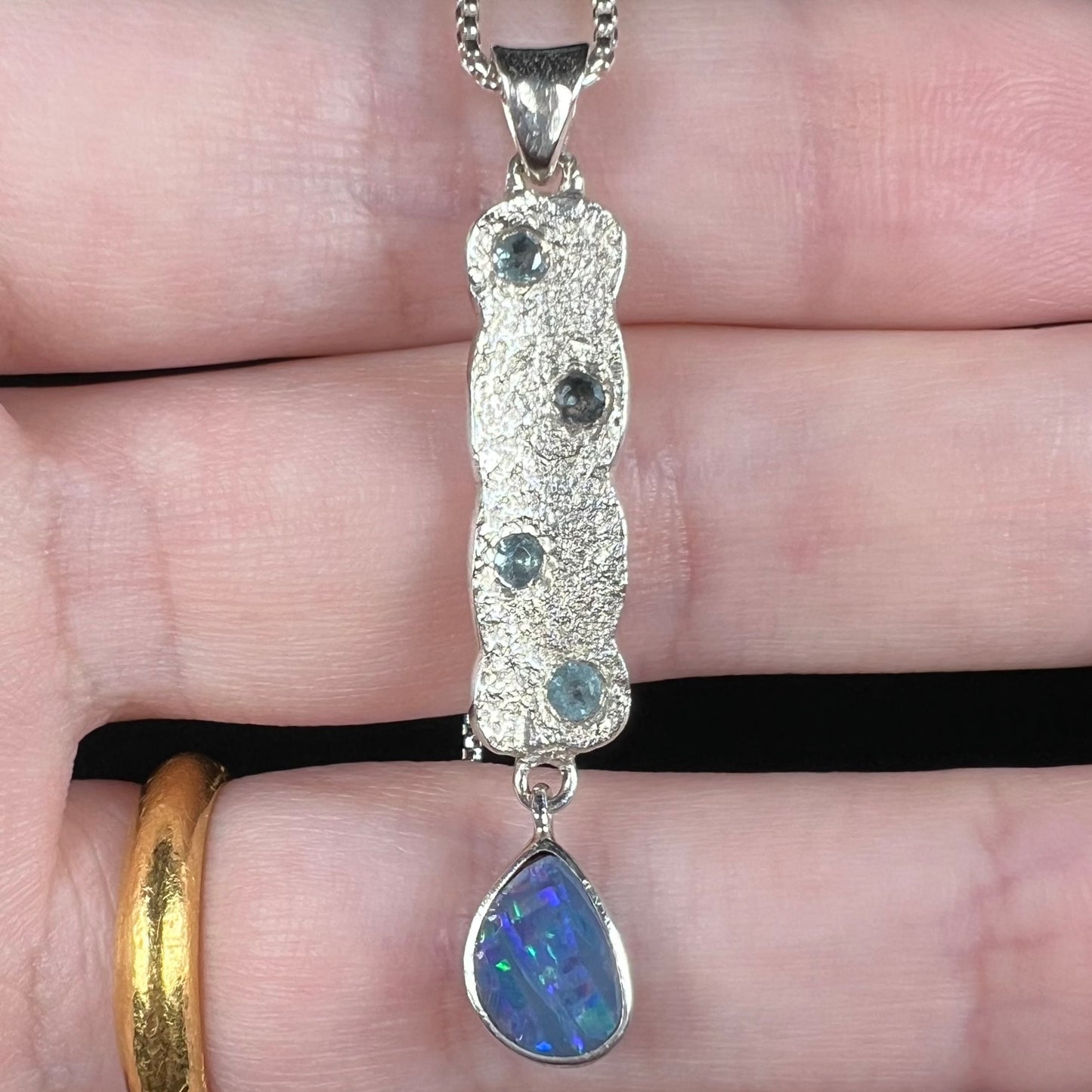 Close-up of an estate Australian black opal doublet and blue topaz drop necklace with a textured sterling silver finish, shown resting on fingers.