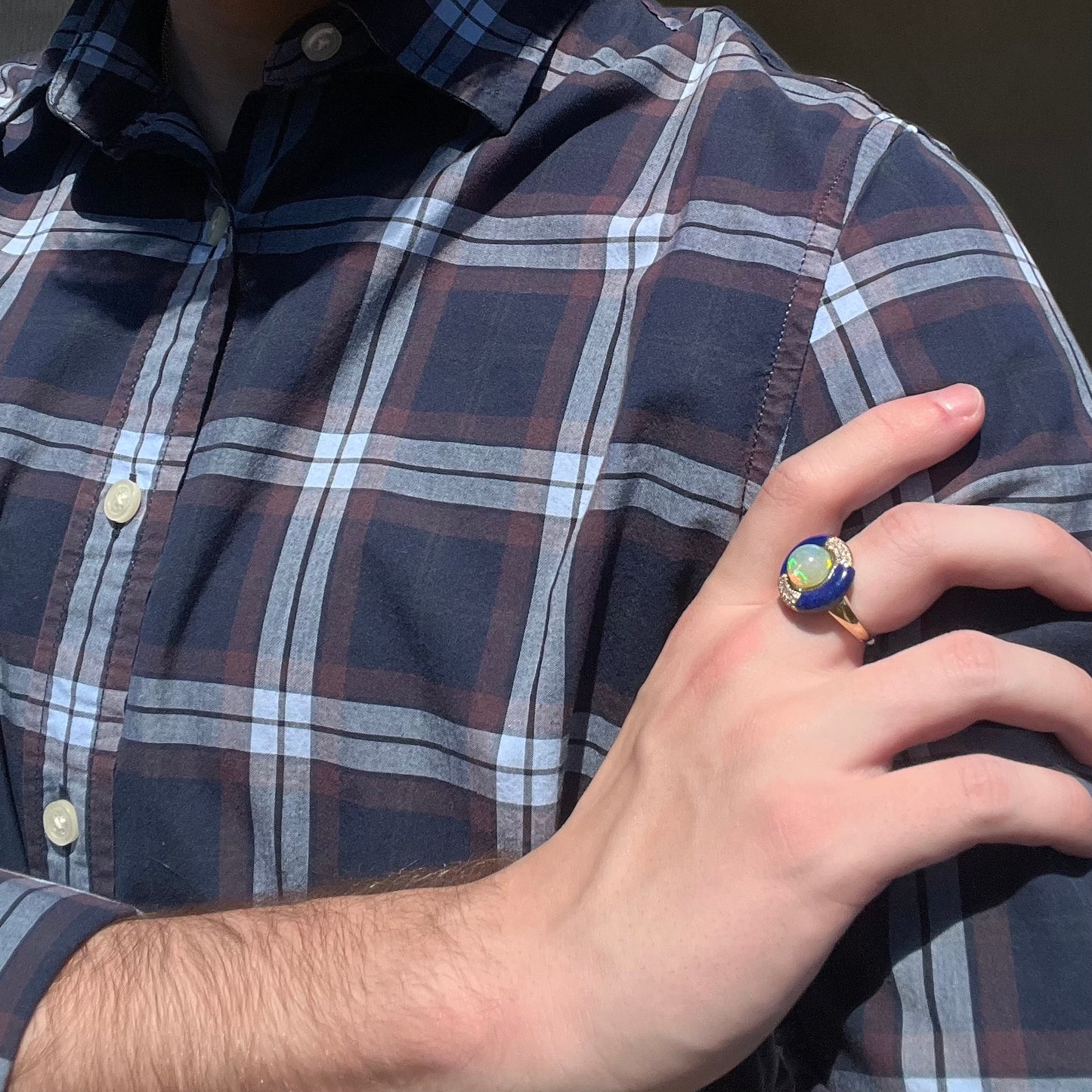 Close-up of a person resting their hand on their opposing shoulder in the sunlight while wearing an estate 14k gold opal, lapis lazuli, and diamond ring.