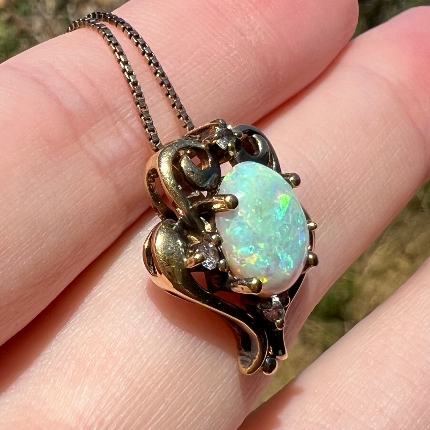 Angled close-up of an estate lab-created opal and CZ pendant in gold-tone metal, shown on fingers in the sunlight.