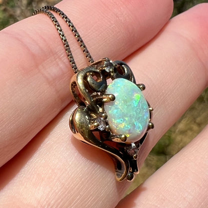 Angled close-up of an estate lab-created opal and CZ pendant in gold-tone metal, shown on fingers in the sunlight.