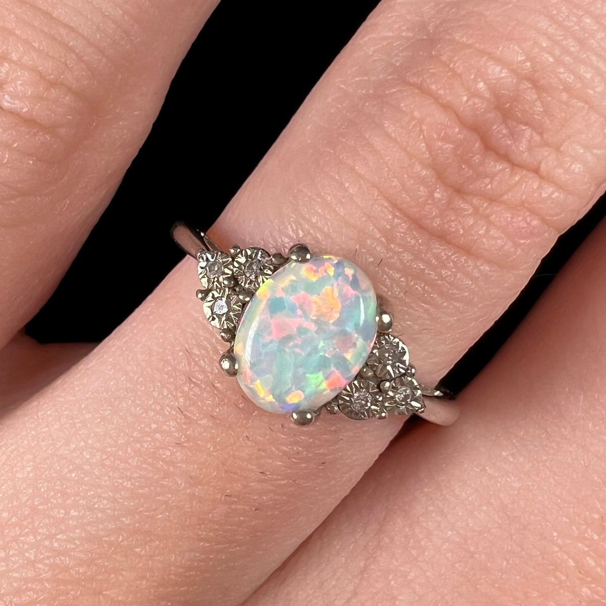 Close-up of an estate sterling silver opal and diamond engagement ring, worn on a finger under studio light.  The opal has multicolor iridescence.