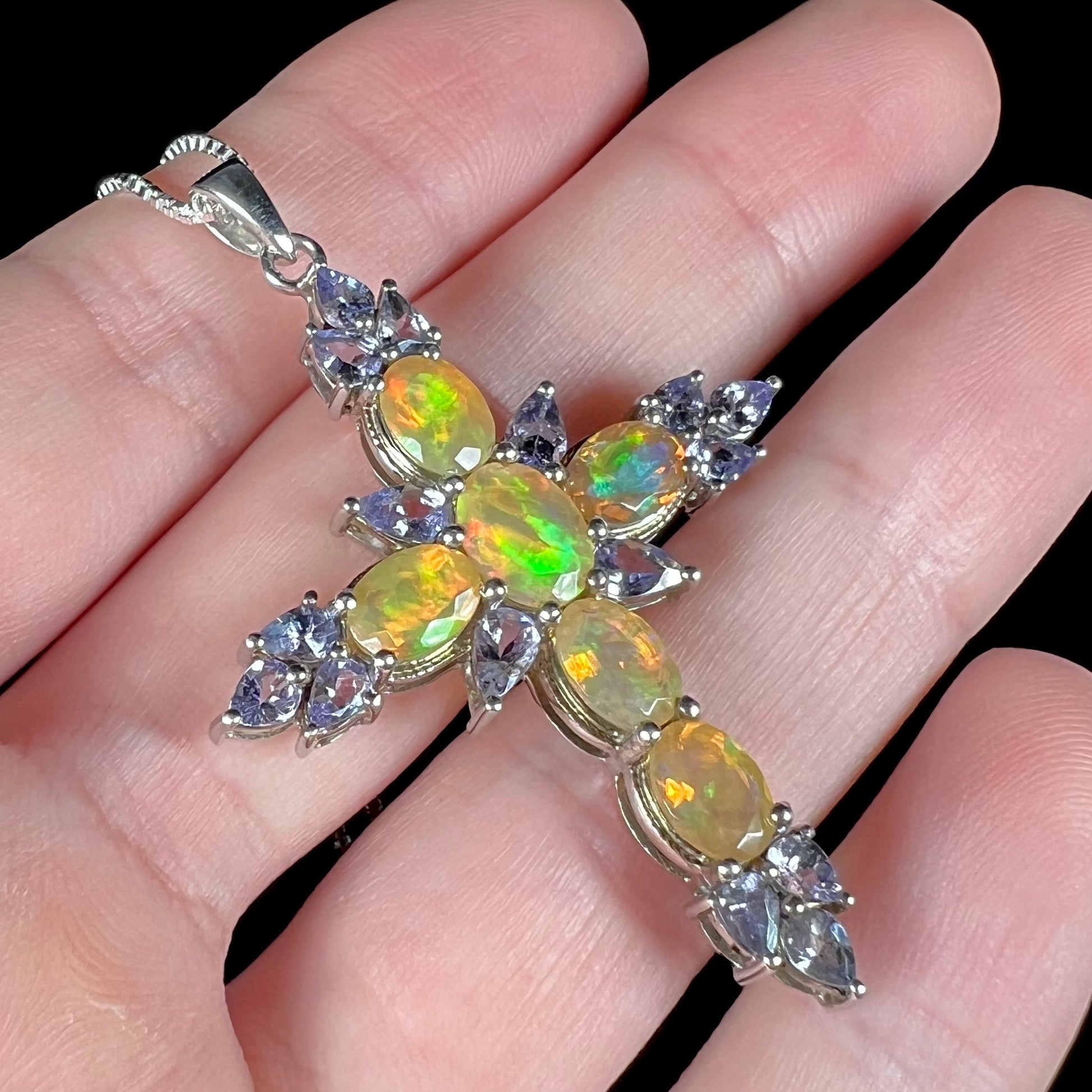 Angled close-up view of a hand holding an Ethiopian fire opal cross necklace with tanzanite accents.