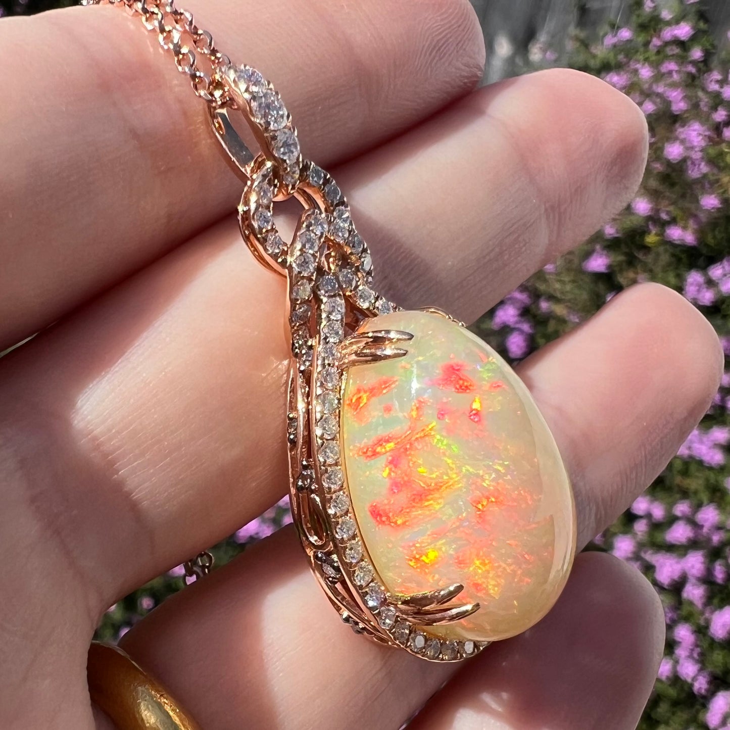 Angled view of an Ethiopian opal and diamond halo pendant resting on a hand in sunlight against a blurred floral background.  The opal has red fire.