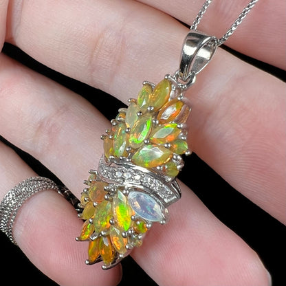 Green flashes seen across faceted marquise and round cut Ethiopian opals mounted in a sterling silver cluster pendant.  Shown held in fingers close-up from angle.