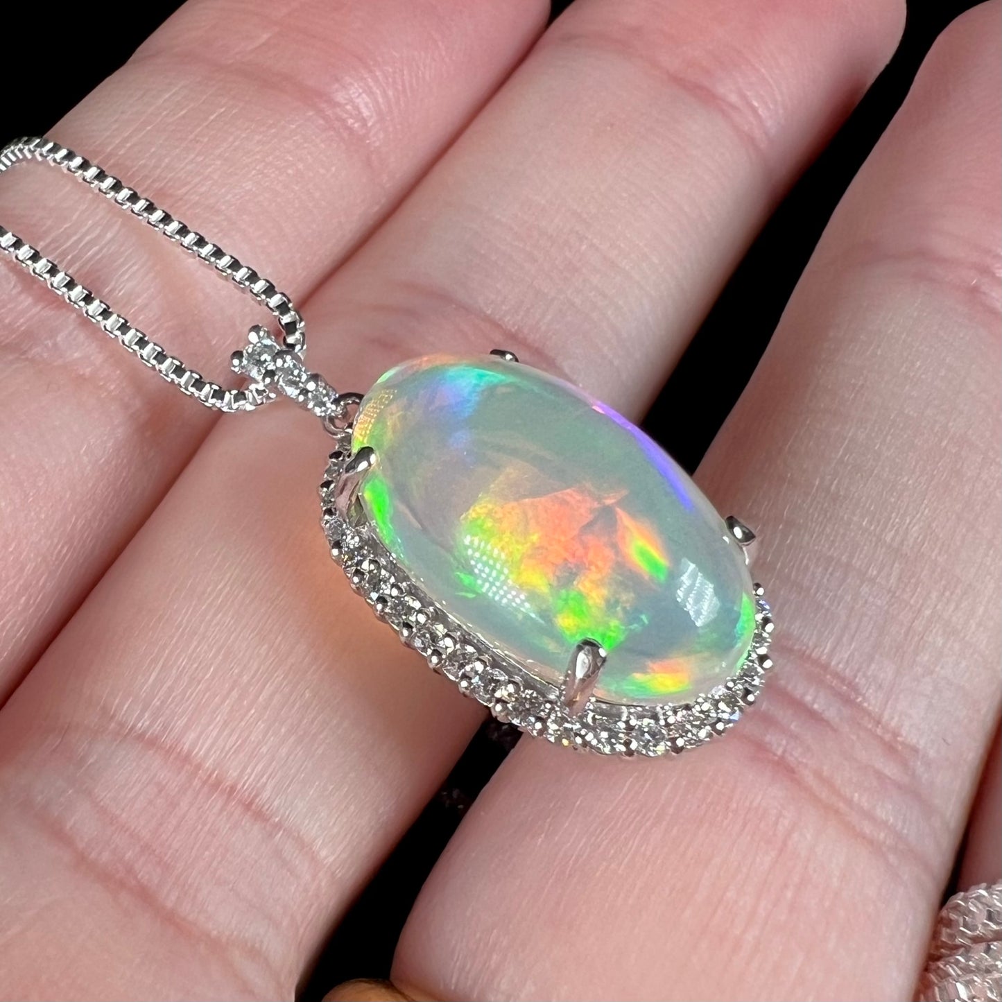 Hand holding an Ethiopian jelly opal and diamond halo pendant from an angle under LED studio light.  The opal has green, red, and yellow play of color.