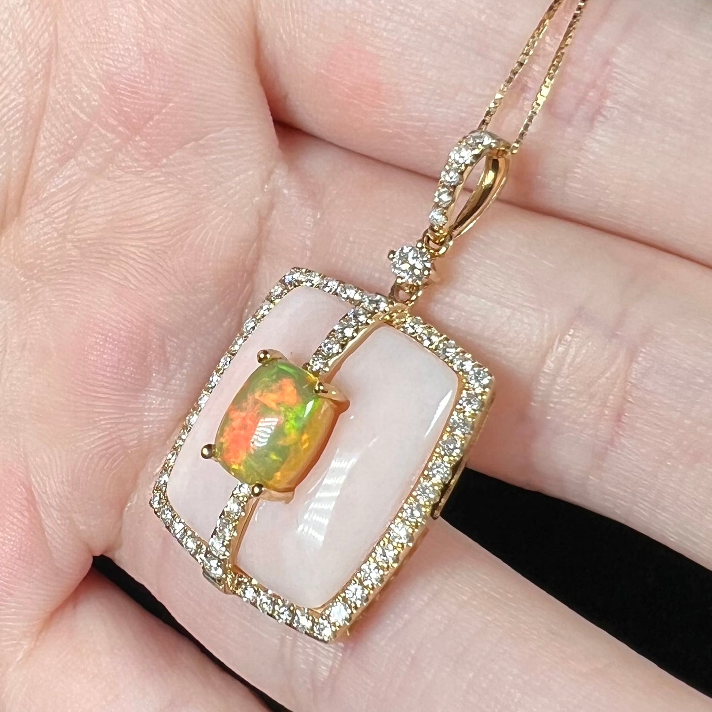 An Ethiopian opal, pink Peruvian opal, and diamond halo pendant in 14k gold shown close-up from angle in hand.