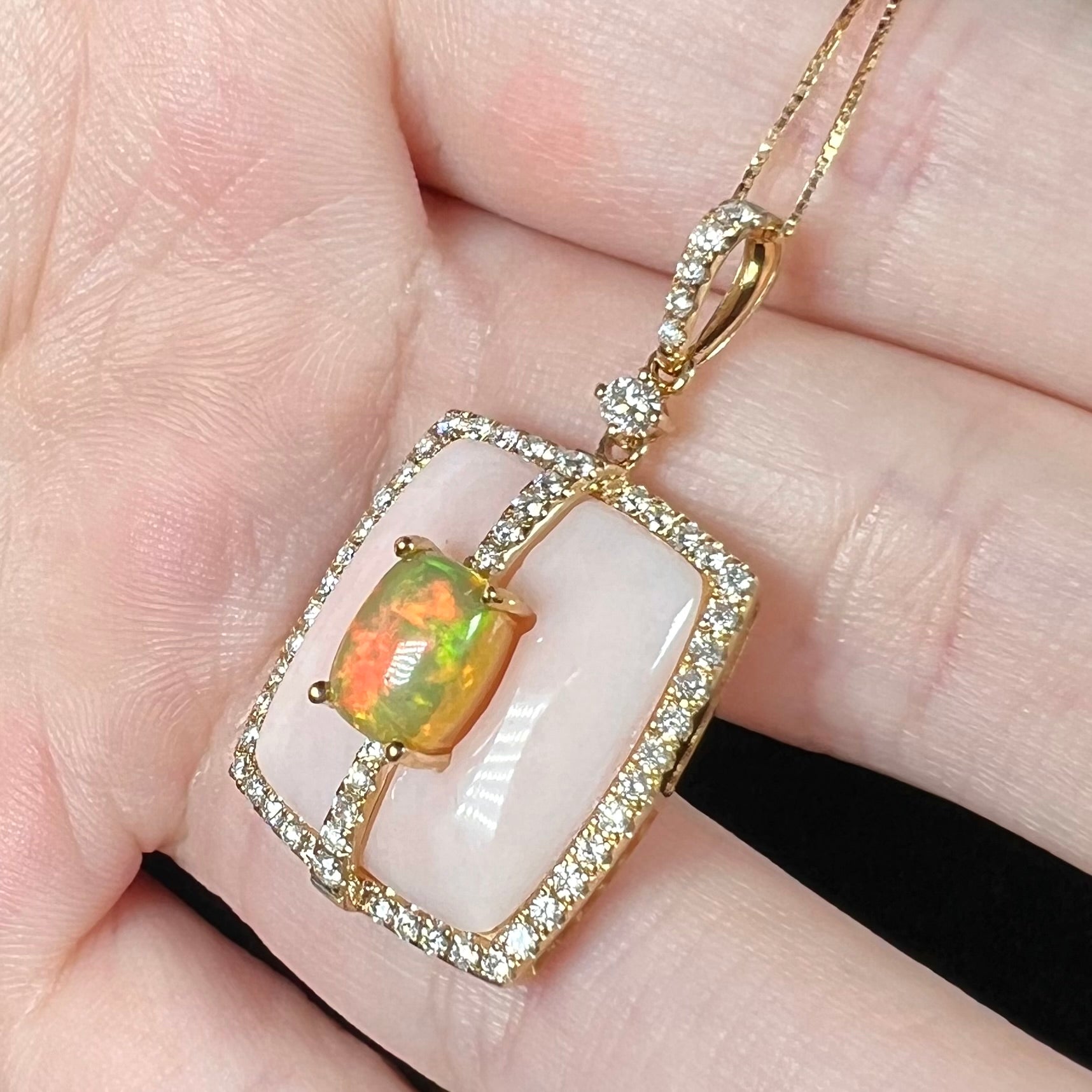 An Ethiopian opal, pink Peruvian opal, and diamond halo pendant in 14k gold shown close-up from angle in hand.