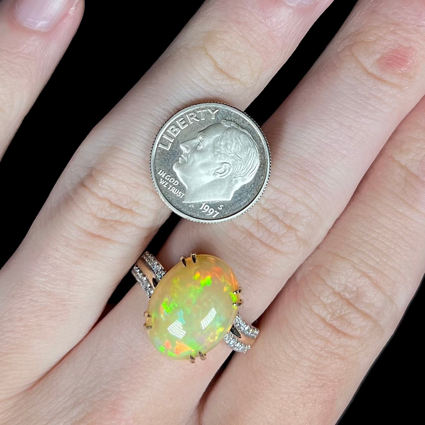 Close-up of hand wearing Ethiopian opal and diamond cocktail ring next to US dime coin for scale; the two are comparable in size