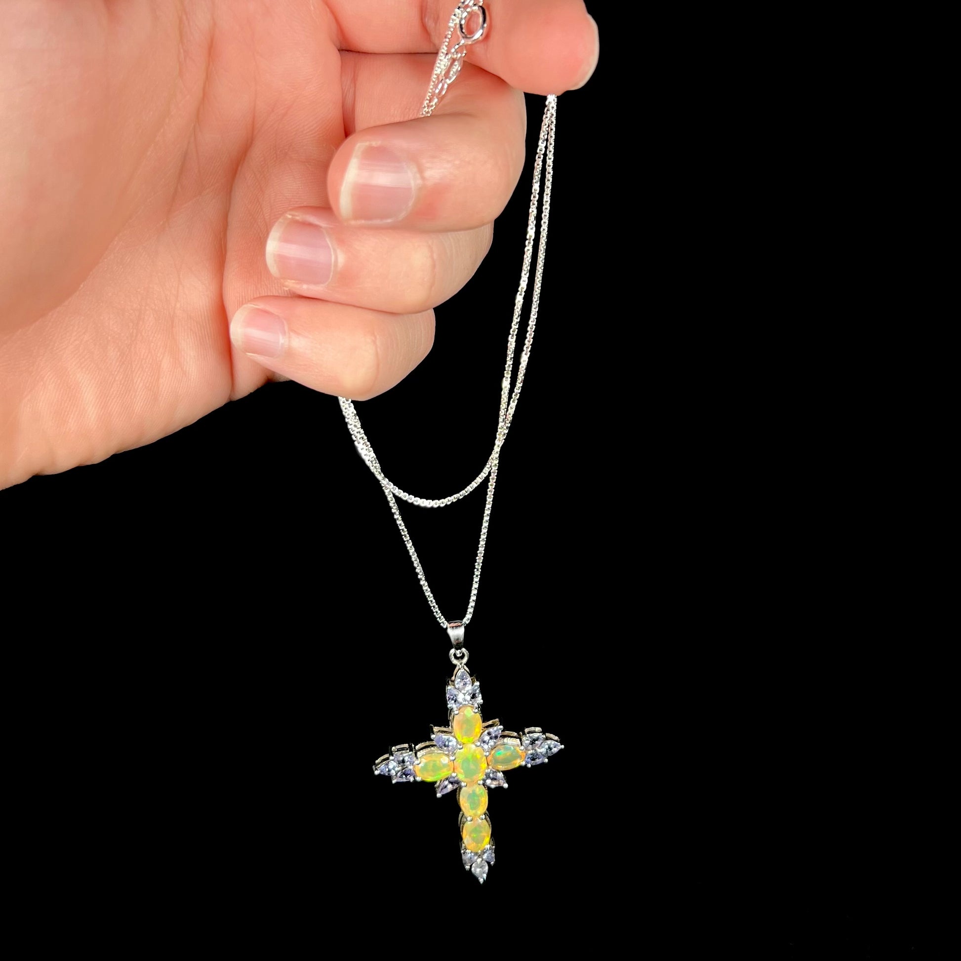 A silver Ethiopian opal cross necklace with tanzanite accents, dangled by a hand from its box-style chain.