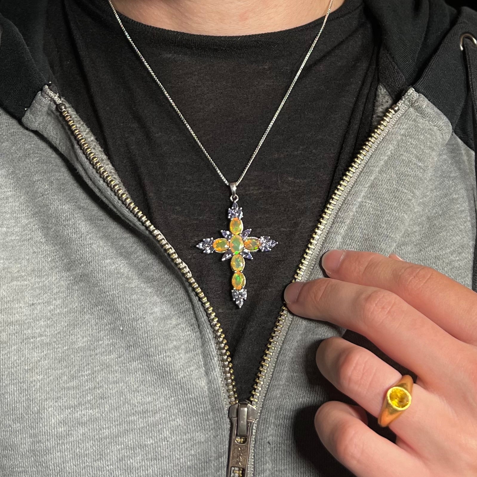 A sterling silver Ethiopian opal cross necklace with tanzanite accents, worn against a dark shirt under an unzipped hoodie.