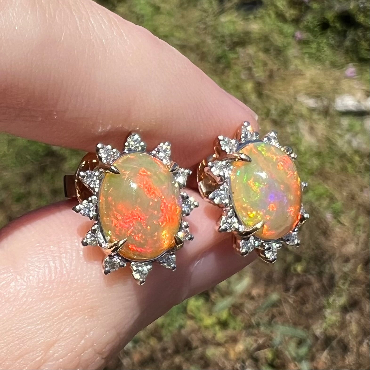 Close-up of Ethiopian opal and diamond stud earrings in 14k gold, held in sunlight between fingers