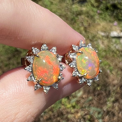 Close-up of Ethiopian opal and diamond stud earrings in 14k gold, held in sunlight between fingers