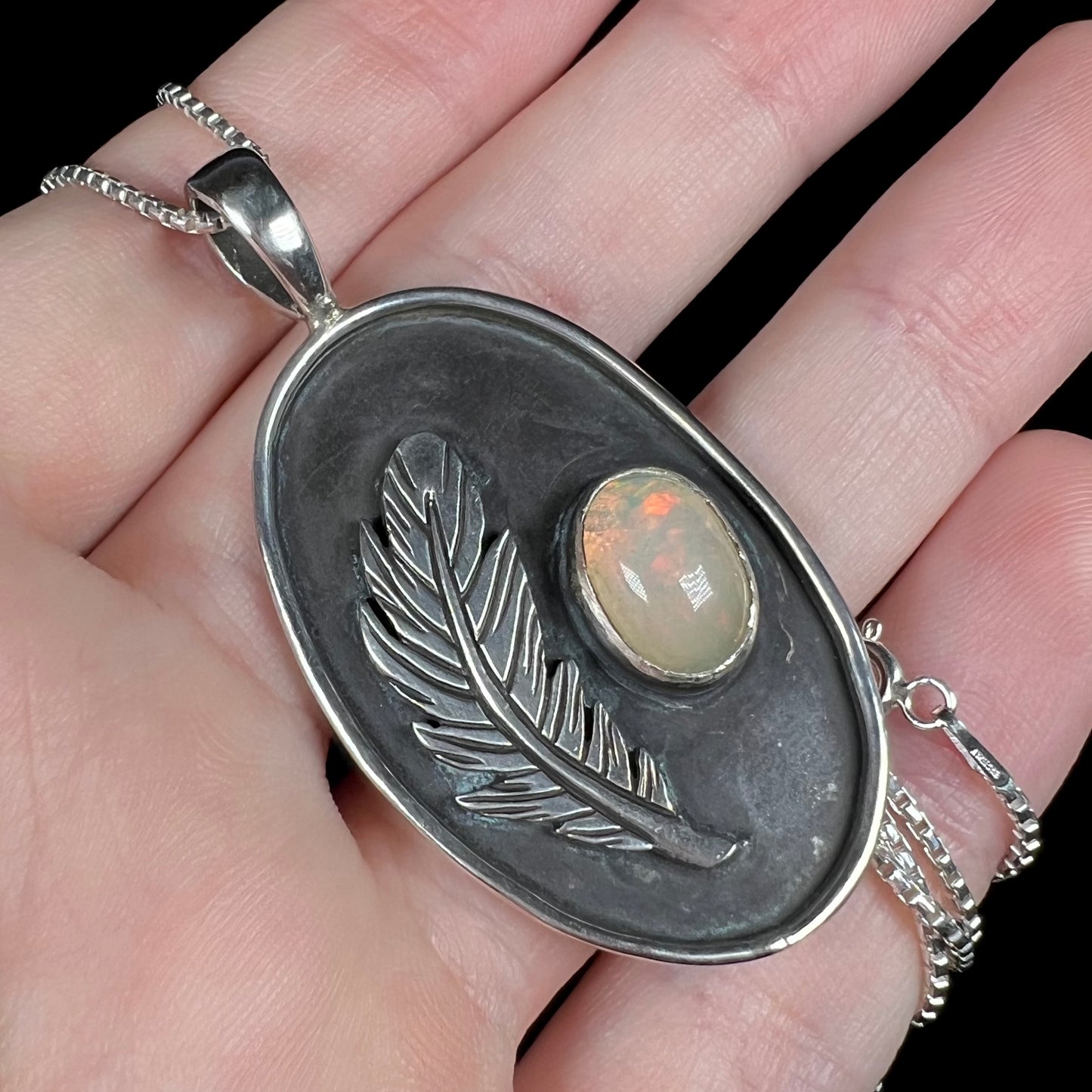Angled close-up of an Ethiopian opal and feather design necklace held in a hand against black backdrop beneath cool LED studio lights.