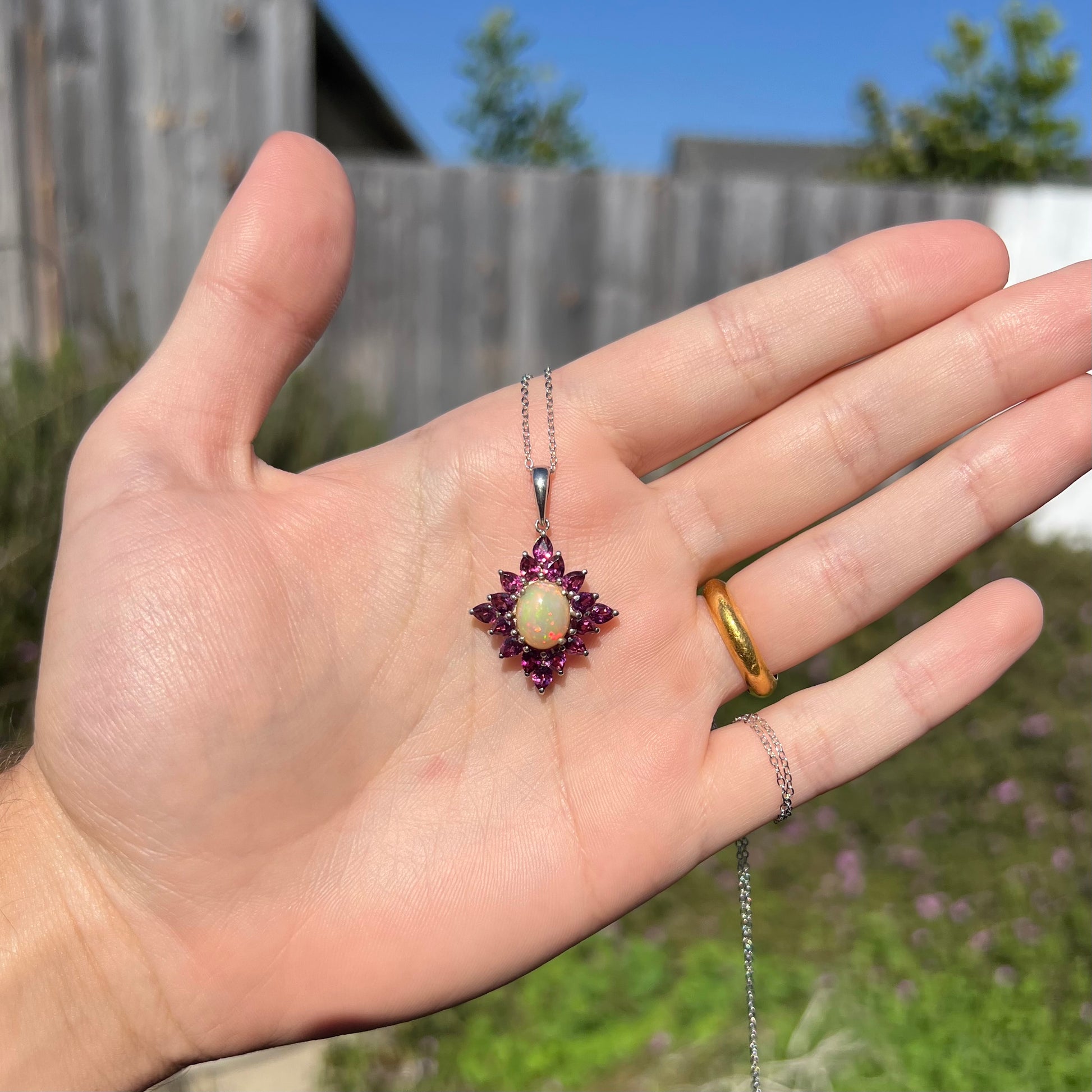 A sterling silver Ethiopian opal and rhodolite garnet spray necklace, shown in a hand in outdoor sunlight.