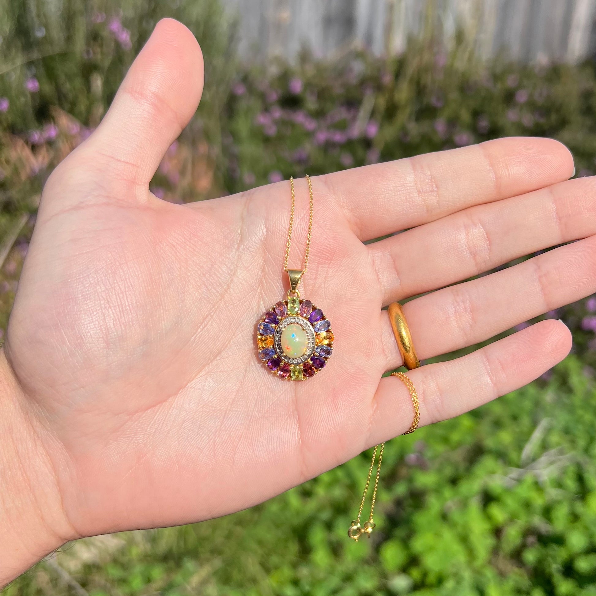 A gold plated sterling silver Ethiopian fire opal and multi-gemstone halo pendant shown in sunlight in a hand.