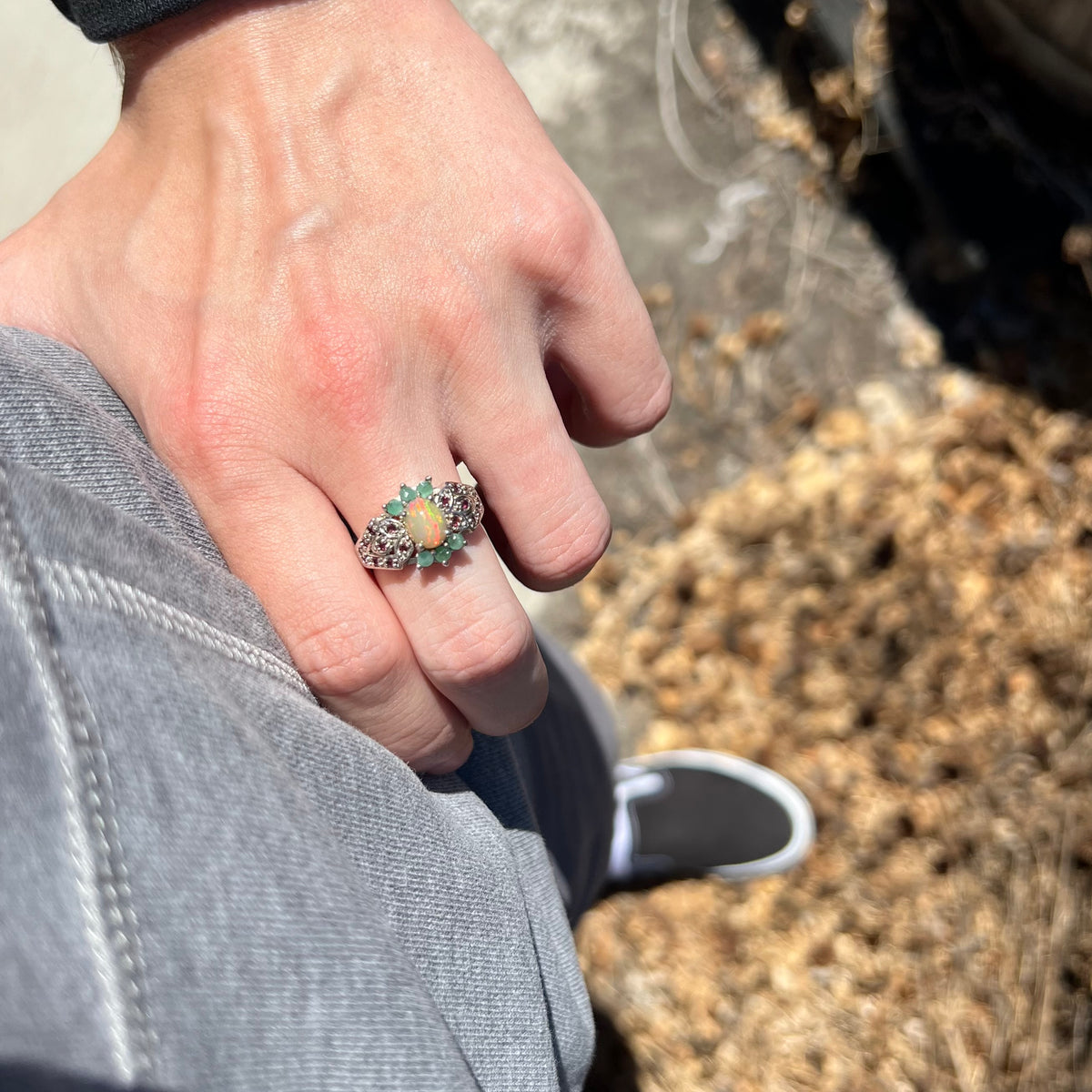 A close-up lifestyle shot of a person's hand resting on hip wearing a silver ring with a central Ethiopian opal and green beryl accents, set against a background of gray denim and dry leaves.
