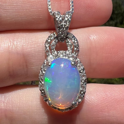 Close-up of women's Ethiopian opal and white topaz halo pendant in sterling silver, held in hand under sunlight