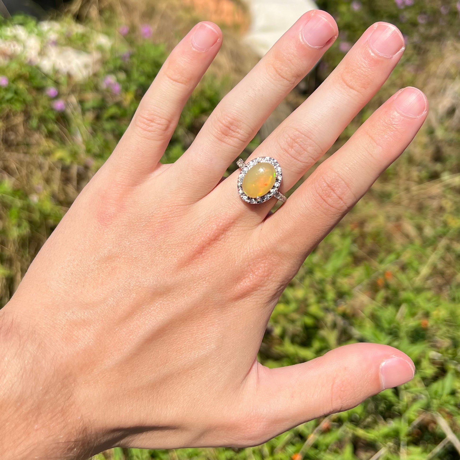 A sterling silver Ethiopian fire opal ring with a CZ halo worn on a hand in the sunlight.