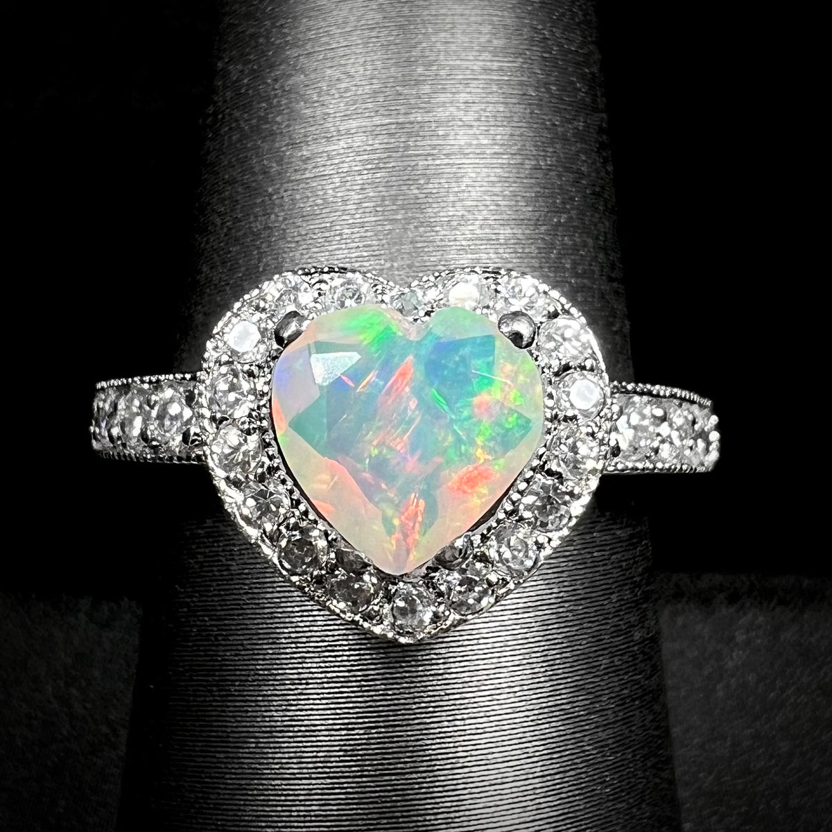 Close-up of heart shaped Ethiopian opal ring with CZ-set halo design and milgrain accents, shown on display