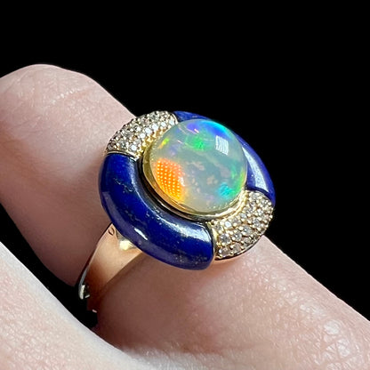 Close-up of an estate designer Ethiopian opal, lapis lazuli, and diamond ring, worn on a finger and shown from an angle under cool studio LED light.