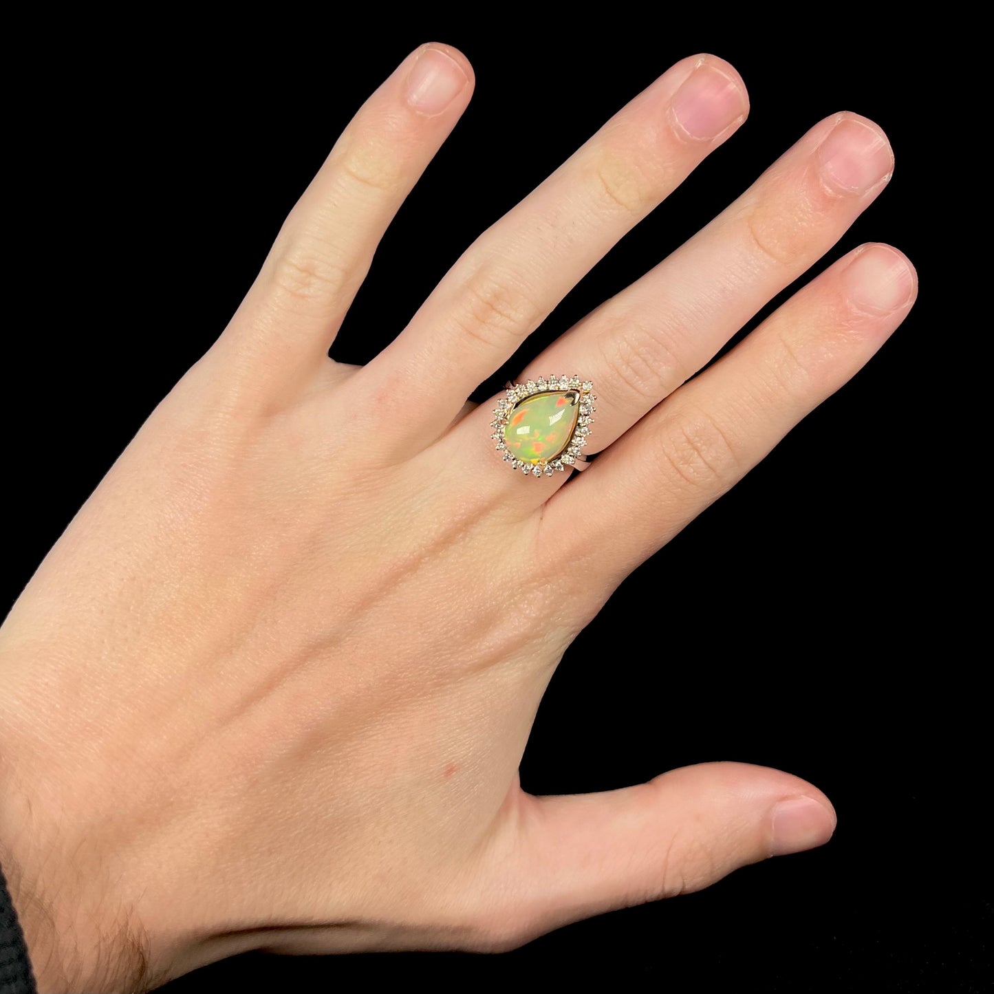 Hand wearing a pear shaped Ethiopian opal and diamond halo engagement ring on middle finger.