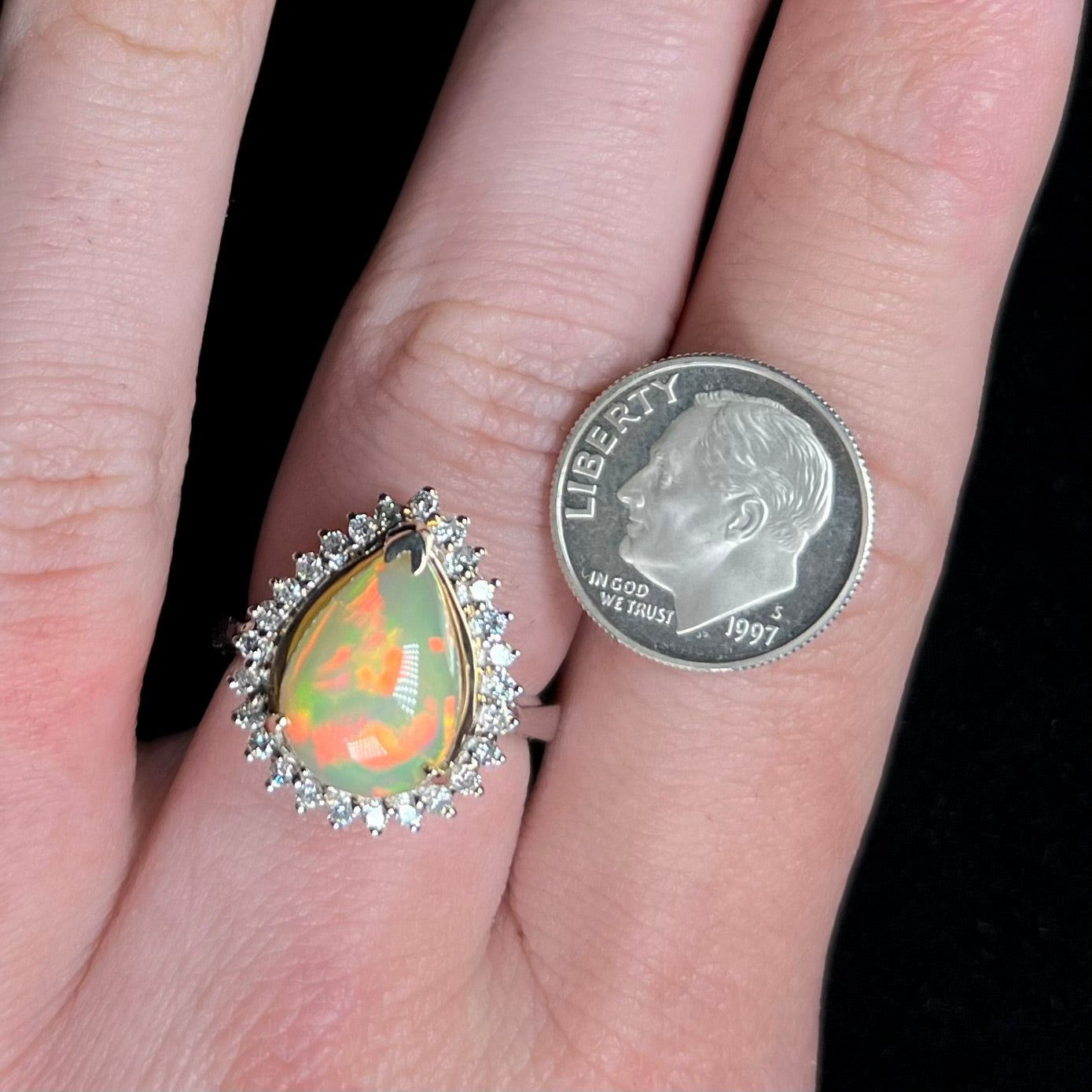 Pear shaped Ethiopian opal and diamond ring with red flashes, worn on a hand next to a US dime coin for scale.
