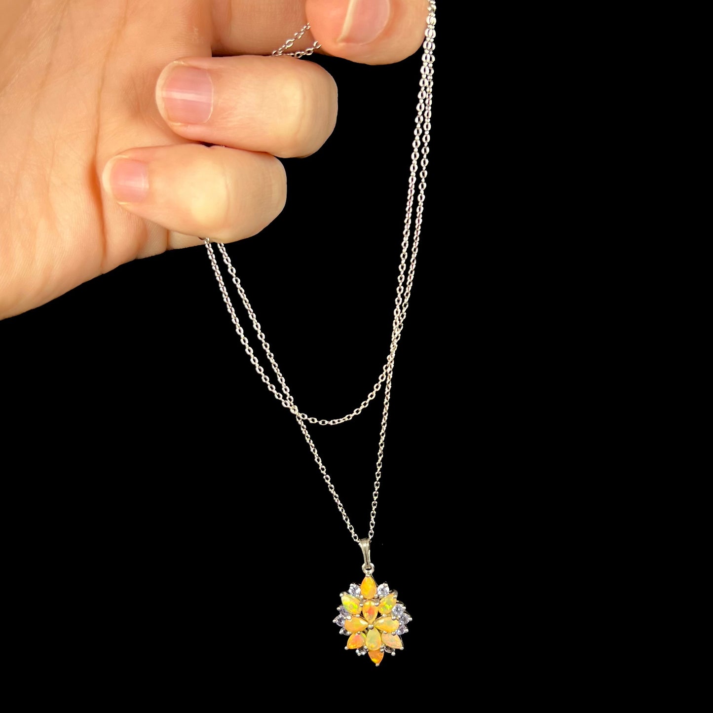 An Ethiopian opal and tanzanite cluster necklace, dangled from its chain by a hand against a black backdrop.