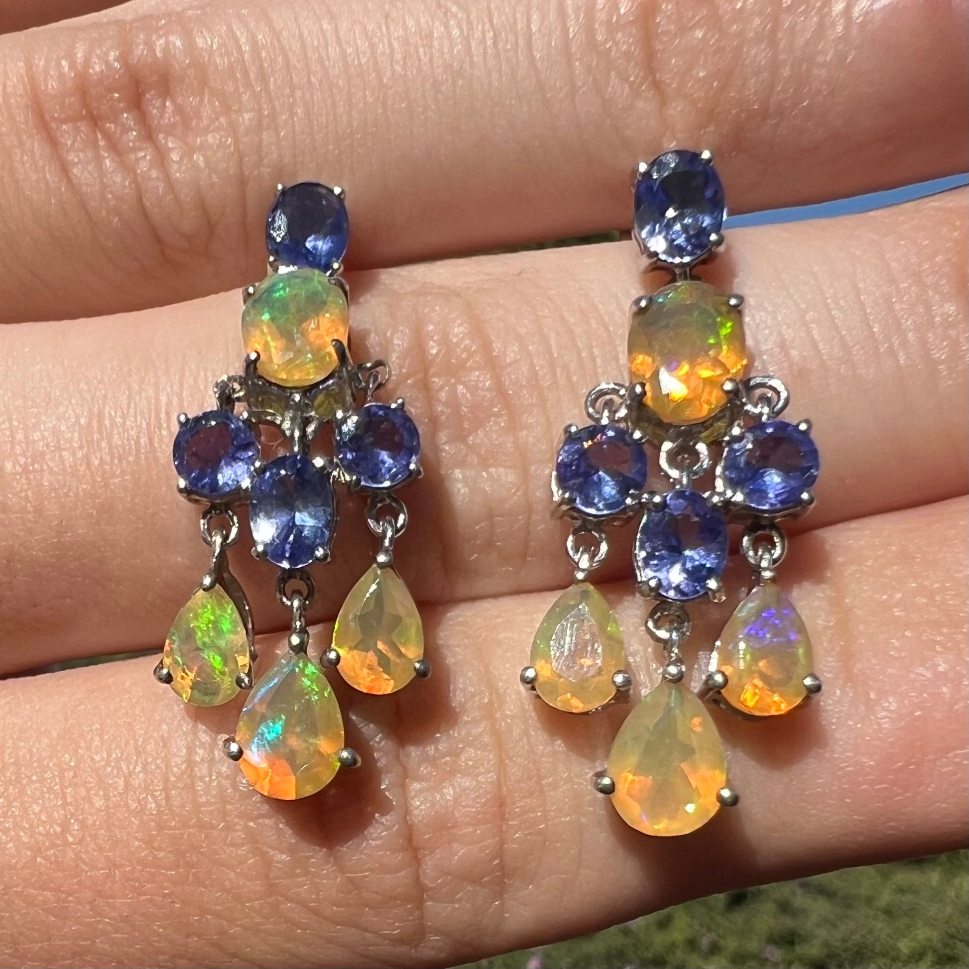 A pair of dangling sterling silver Ethiopian opal and tanzanite chandelier earrings, close-up in sunlight.