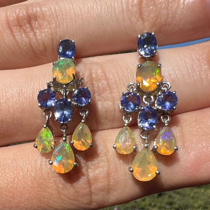 A pair of dangling sterling silver Ethiopian opal and tanzanite chandelier earrings, close-up in sunlight.