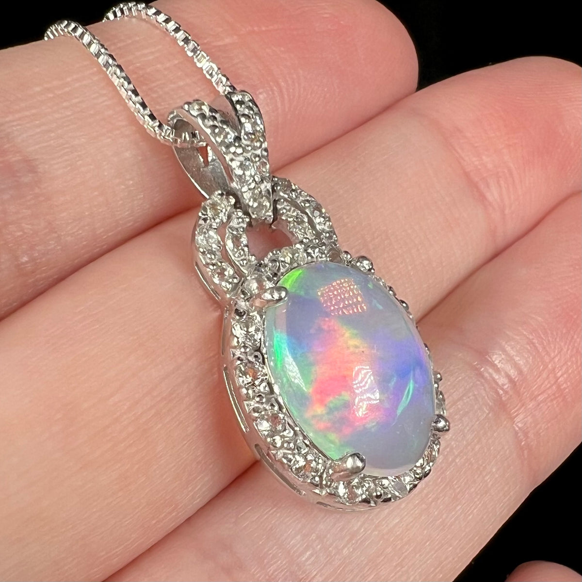Angled close-up of Ethiopian opal and white topaz halo pendant in sterling silver, held in hand