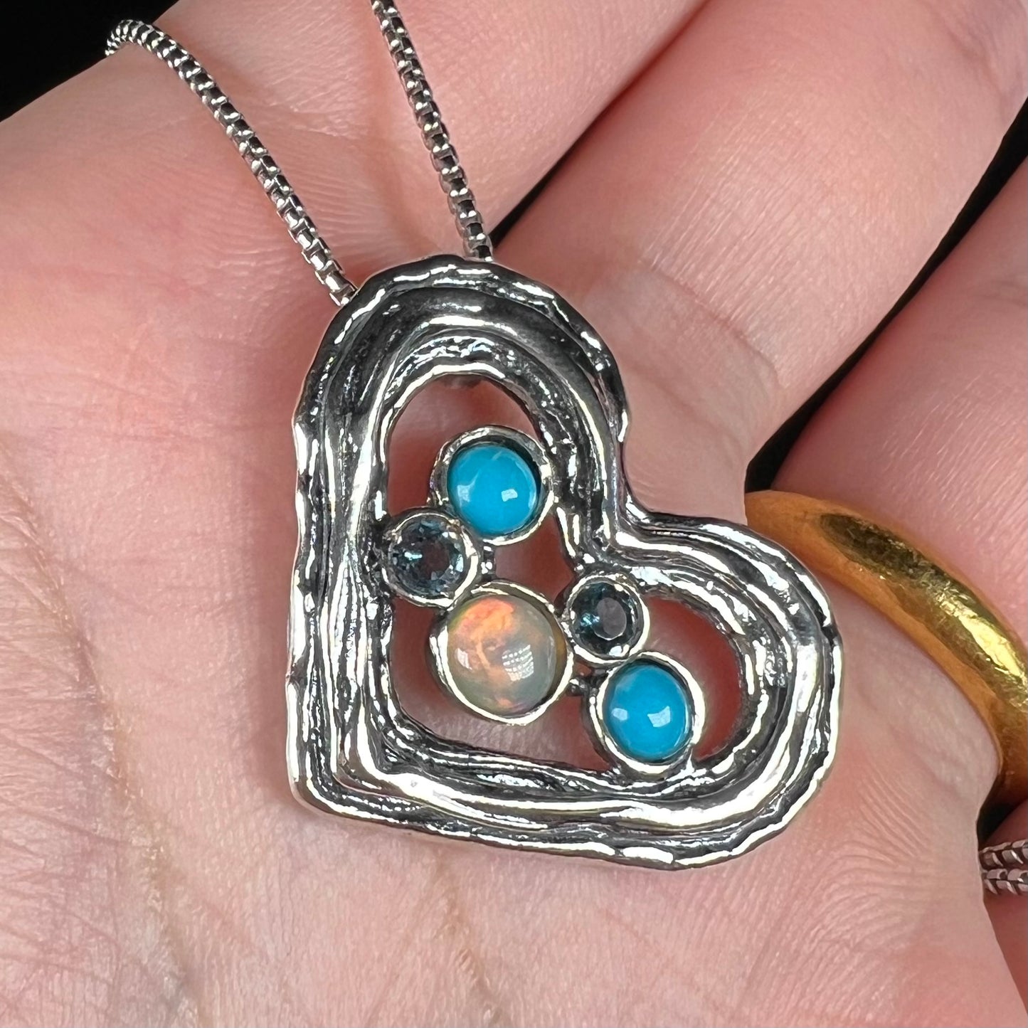 Angled close-up of a sterling silver heart necklace set with opal, turquoise, and blue topaz in a hand.