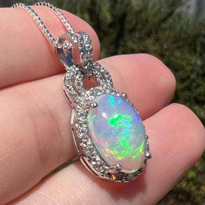 Angled close-up of Ethiopian opal and white topaz halo pendant held in sunlight.  Opal has green flashes of color