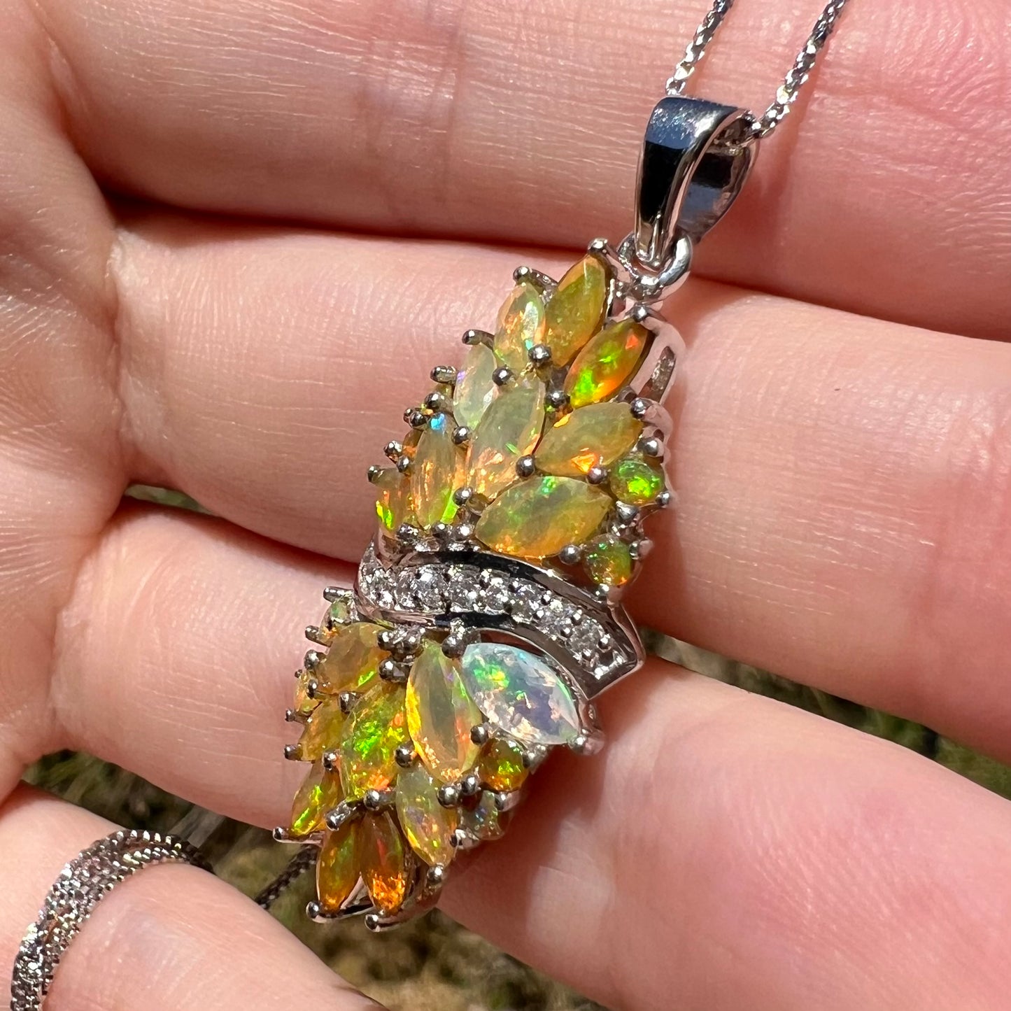 Macro close-up showing sterling silver Ethiopian opal pendant with white zircon accents in sunlight, held in a hand.  Green flashes are seen in the table facets of the faceted opals.