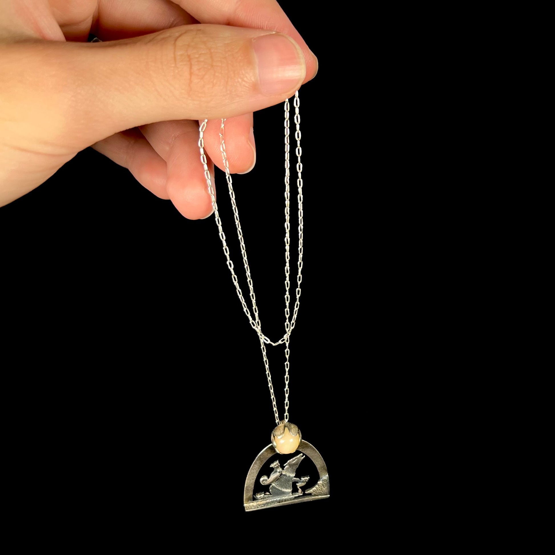 A sterling silver Ethiopian opal wolf and cat design necklace, dangled from a hand by its chain against black.