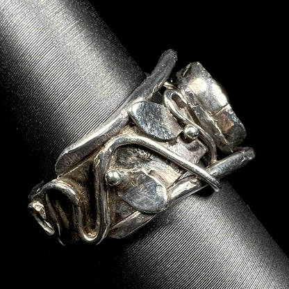Hand-fabricated sterling silver ring with intricate scrolling vine and leaf design along shank, viewed on ring display from side