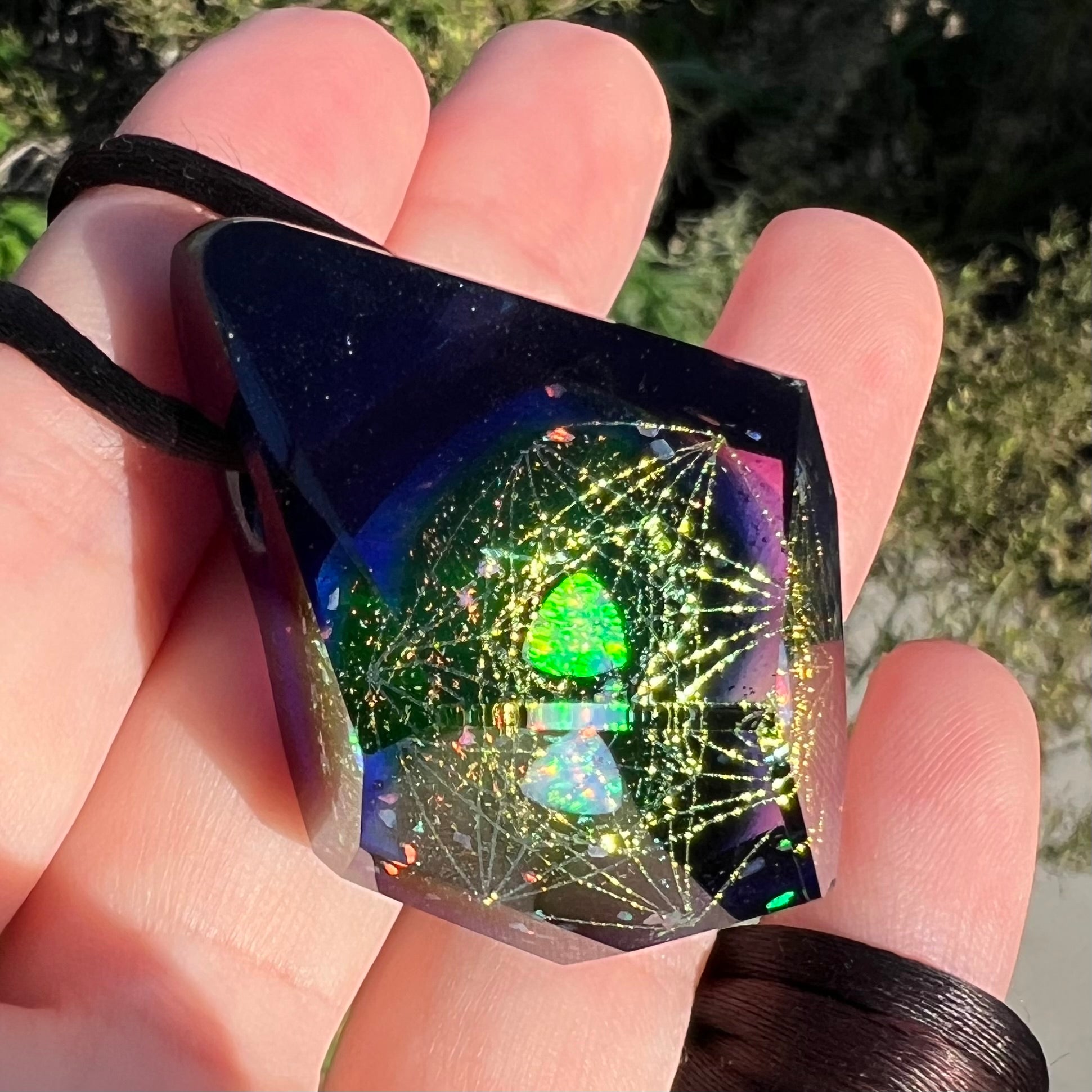A faceted purple borosilicate glass pendant with a green lab-created opal inside, shown close-up from an angle in the sunlight, held in a hand.