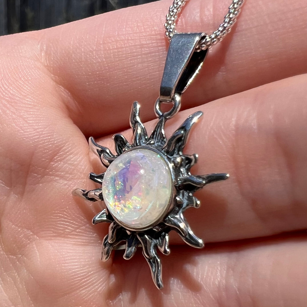 Close-up angled view of a fake, imitation glass opal sunburst pendant in sterling silver on a chain, shown in natural sunlight.