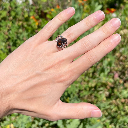 Fire agate ring on hand in sunlight with green bushes in background.