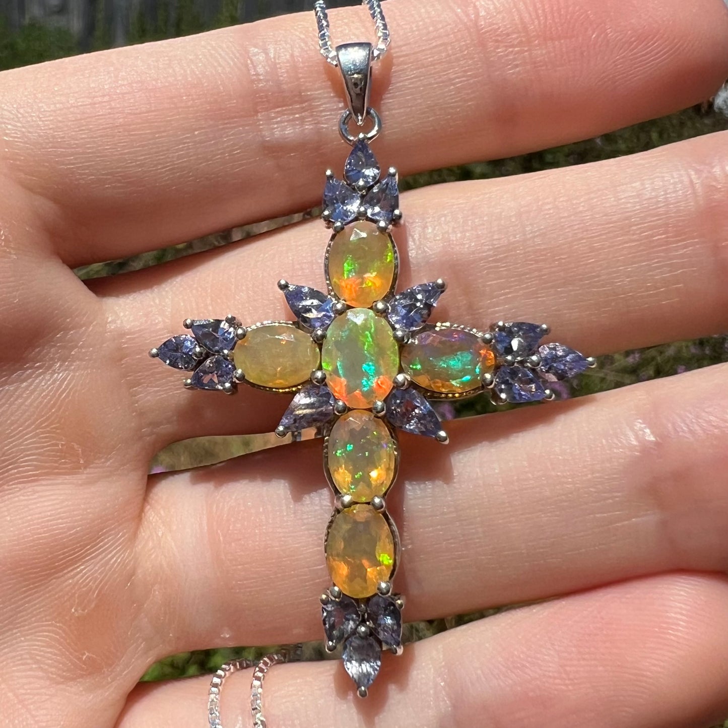 Close-up front view of a fire opal cross necklace with tanzanite stones in the sunlight.