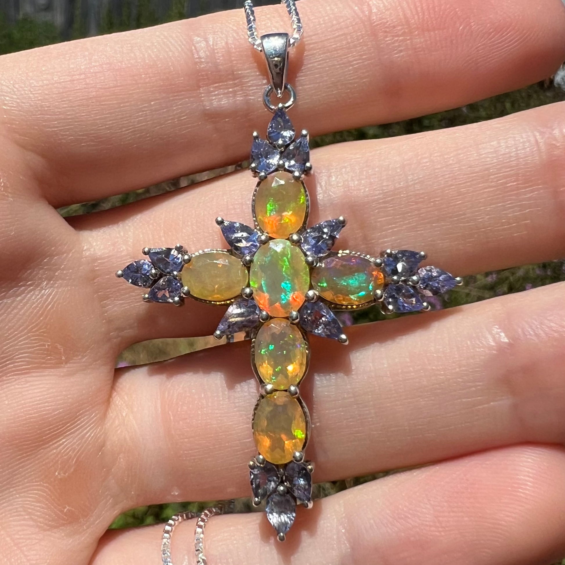Close-up front view of a fire opal cross necklace with tanzanite stones in the sunlight.