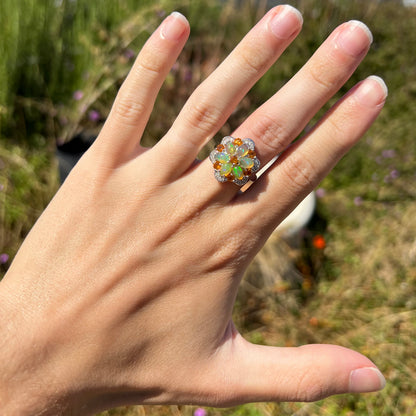 A hand wearing a fire opal and diamond flower-design ring in the sunlight against blurred outdoor background.