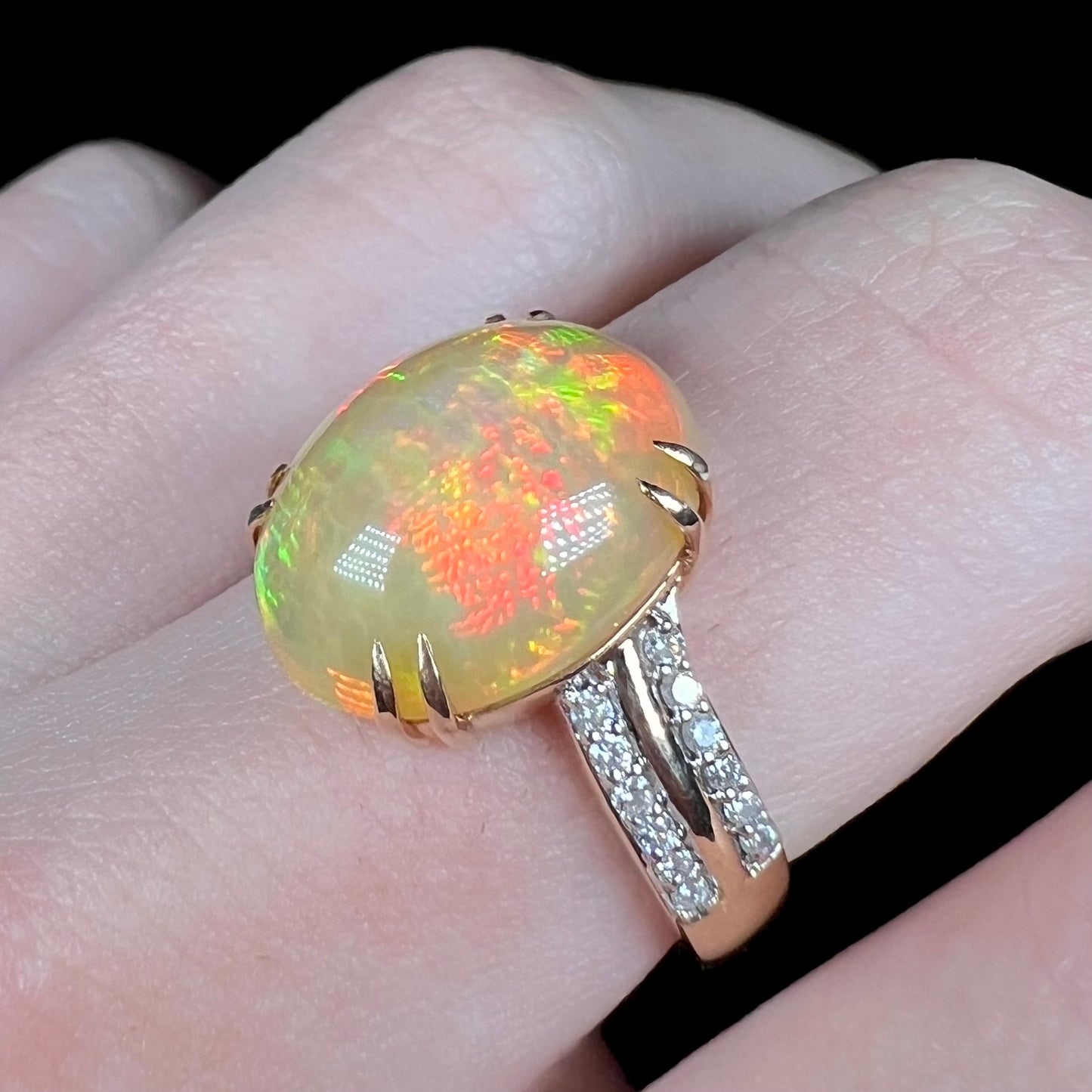 Angled close-up of women's 14k gold fire opal and diamond ring worn on finger.  Opal has bright red and green flashes of color