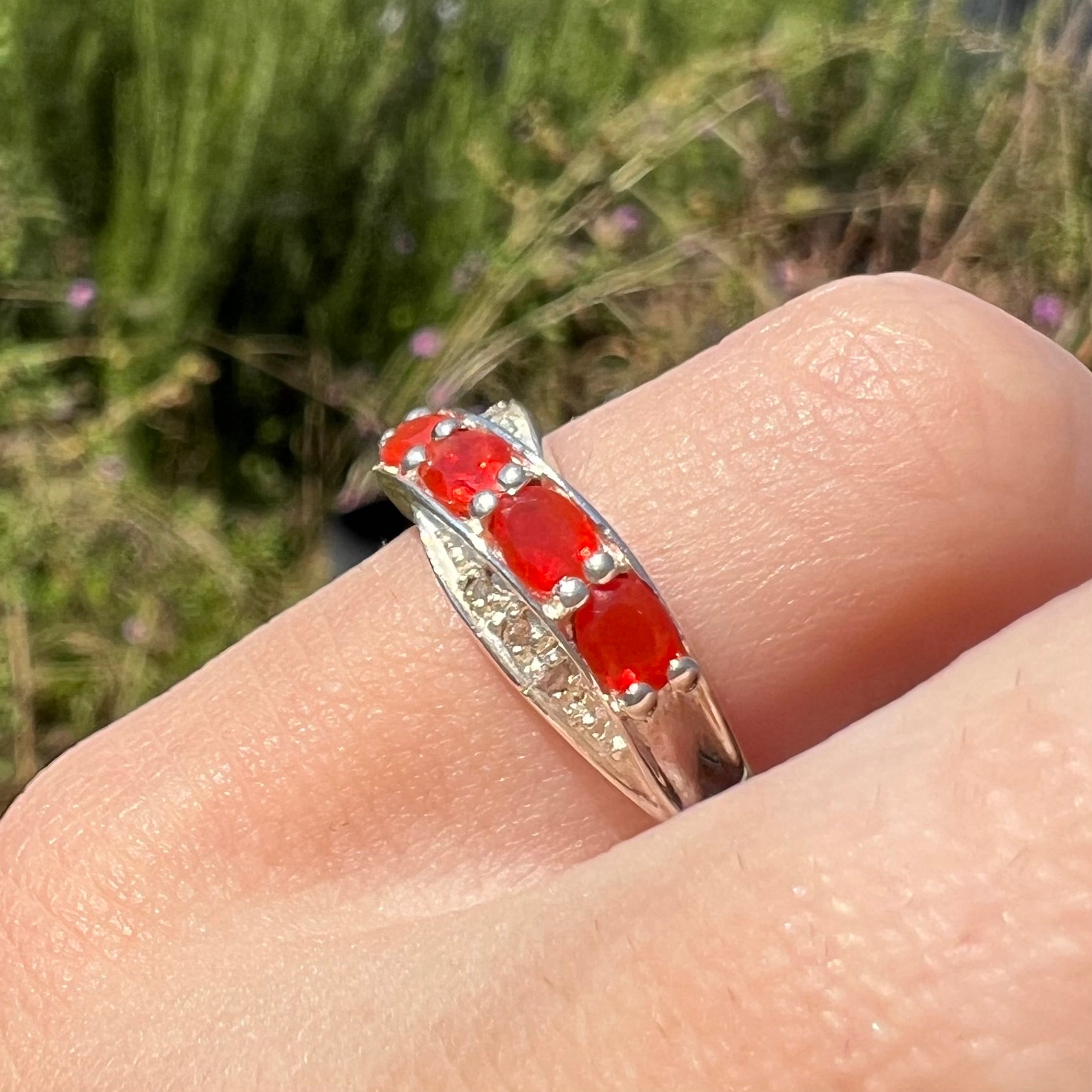 Angled close-up of silver fire opal and diamond ring worn on hand in sunlight against blurred natural background.