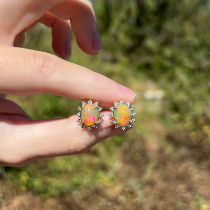 Fingers holding Ethiopian fire opal and diamond halo stud earrings in natural sunlight