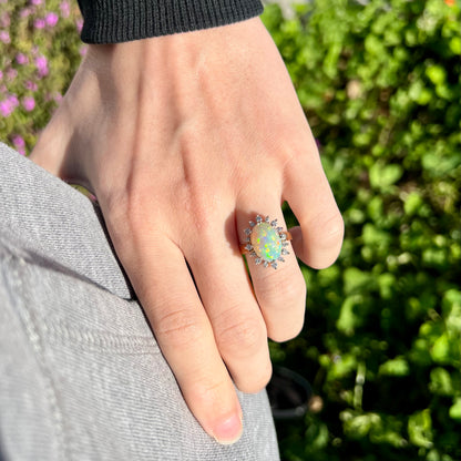 A lifestyle shot of a 4.30ct Lightning Ridge fire opal engagement ring worn on a hand resting on the hip in sunlight.  Green plants are visible in the background.