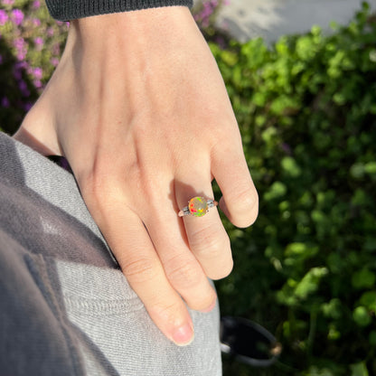 A platinum fire opal and diamond engagement ring worn on a model's hand in the sunlight, showcasing the opal's vivid red and green play-of-color.