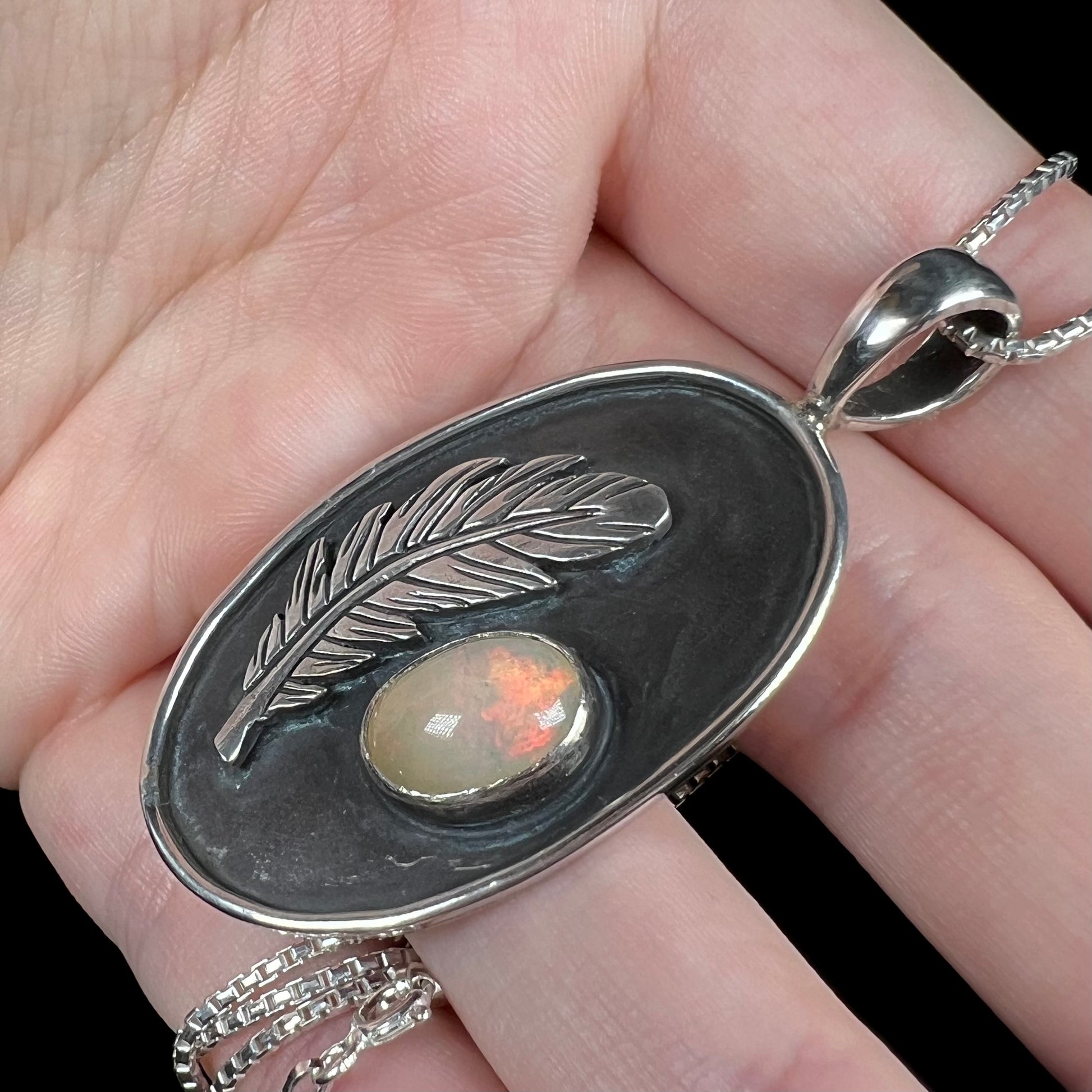 Hand holding a sterling silver fire opal men's necklace in from an angle against black background.  The opal has faint red play of color under cool studio lights.