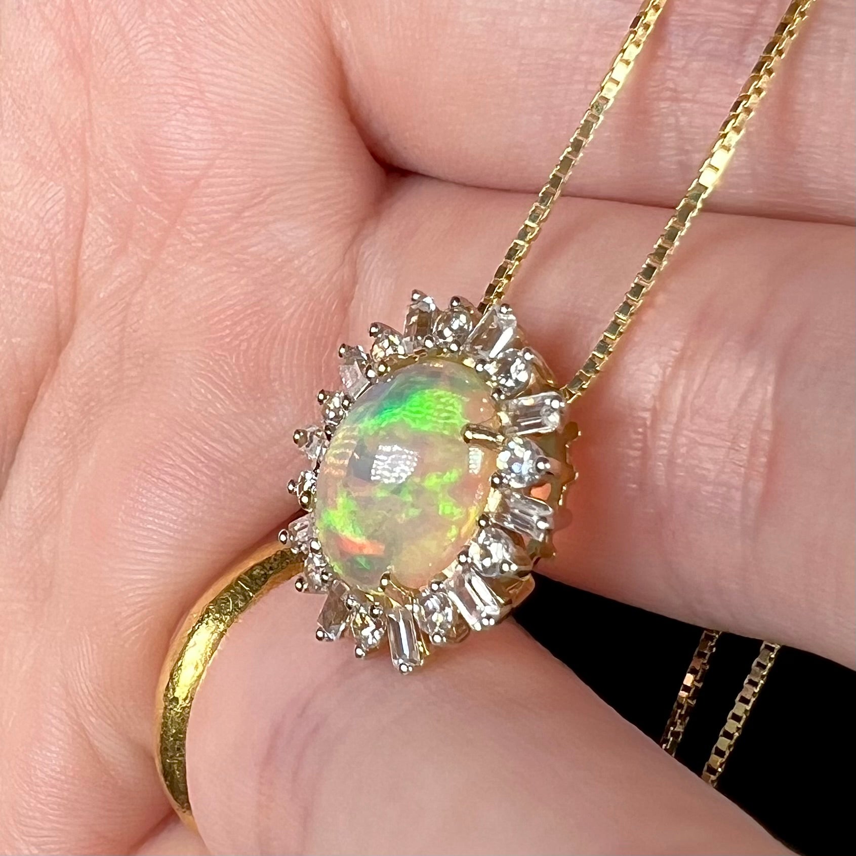 Yellow gold fire opal pendant with spiked halo of emerald cut and round brilliant cut white sapphires, shown from angle close-up in hand.